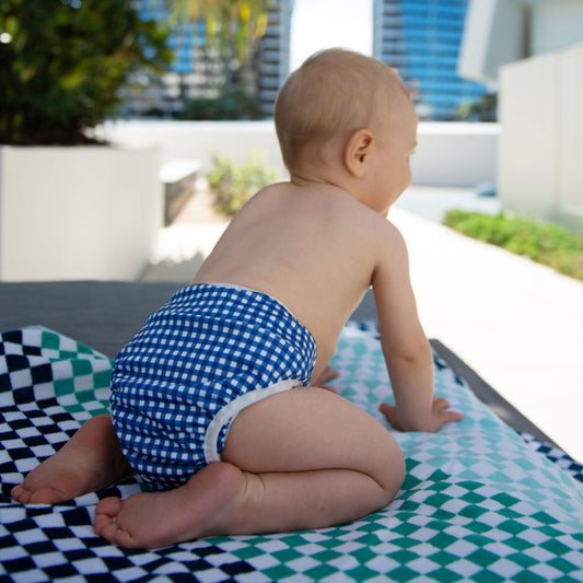 Happy toddler wearing a Tyoub blue check reusable swim nappy by an outdoor pool in Australia.