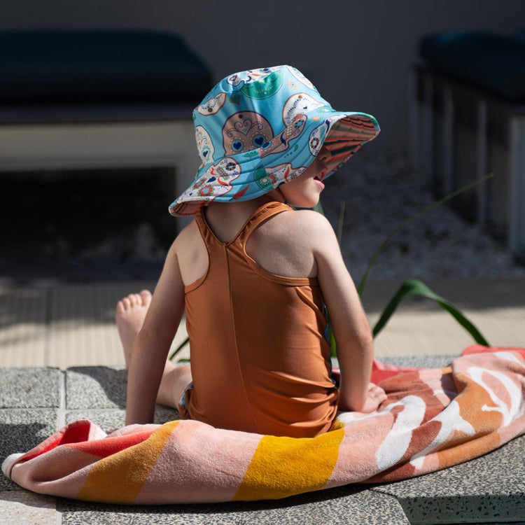 Child wearing the Tyoub sugar skull kneesuit zootie and matching sun hat sitting by an outdoor pool in Australia back view.