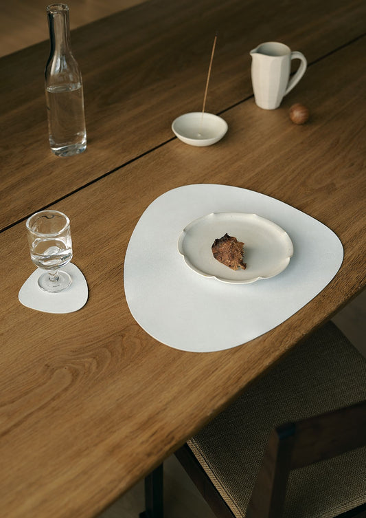 Recycled leather tablemats on a wooden table with a white plate, glass, and small bowl.