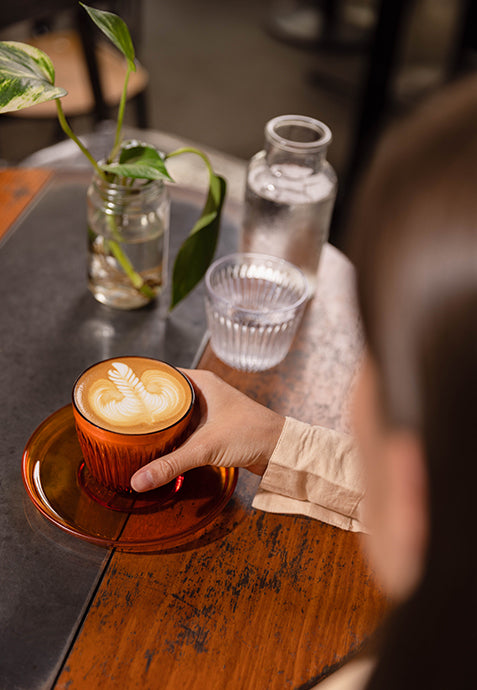Person holding a cup of coffee with latte art on a wooden table with a plant and water bottle in the background.