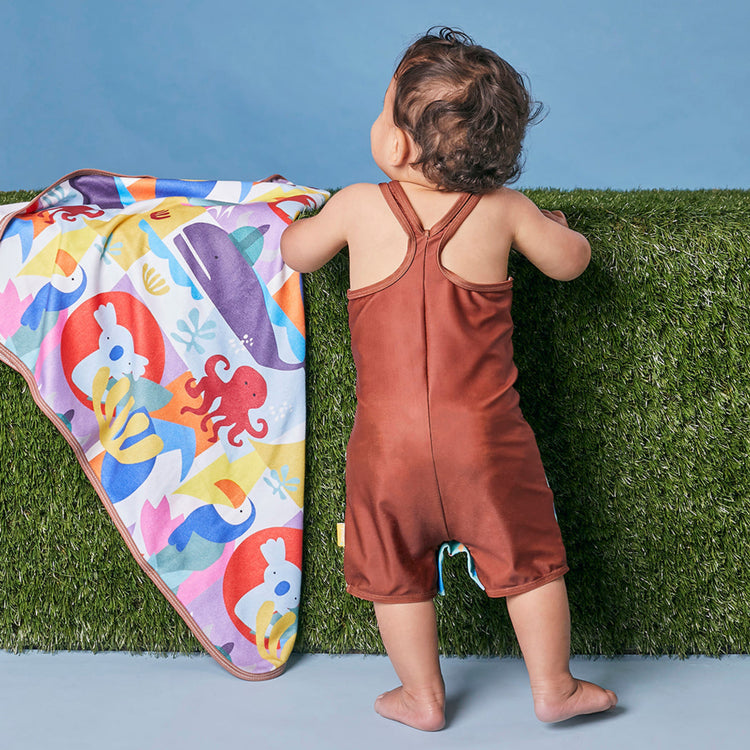 Toddler wearing the Tyoub ocean glass color block zootie swimsuit standing by a colorful baby beach rug showing back.