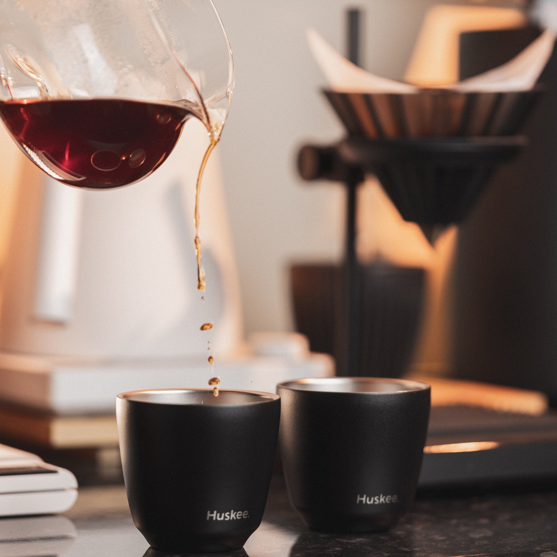 Set of 2 HuskeeSteel 3oz espresso cups crafted from 91% recycled double-wall vacuum insulated stainless steel on a cafe counter with coffee being poured from a glass jug.