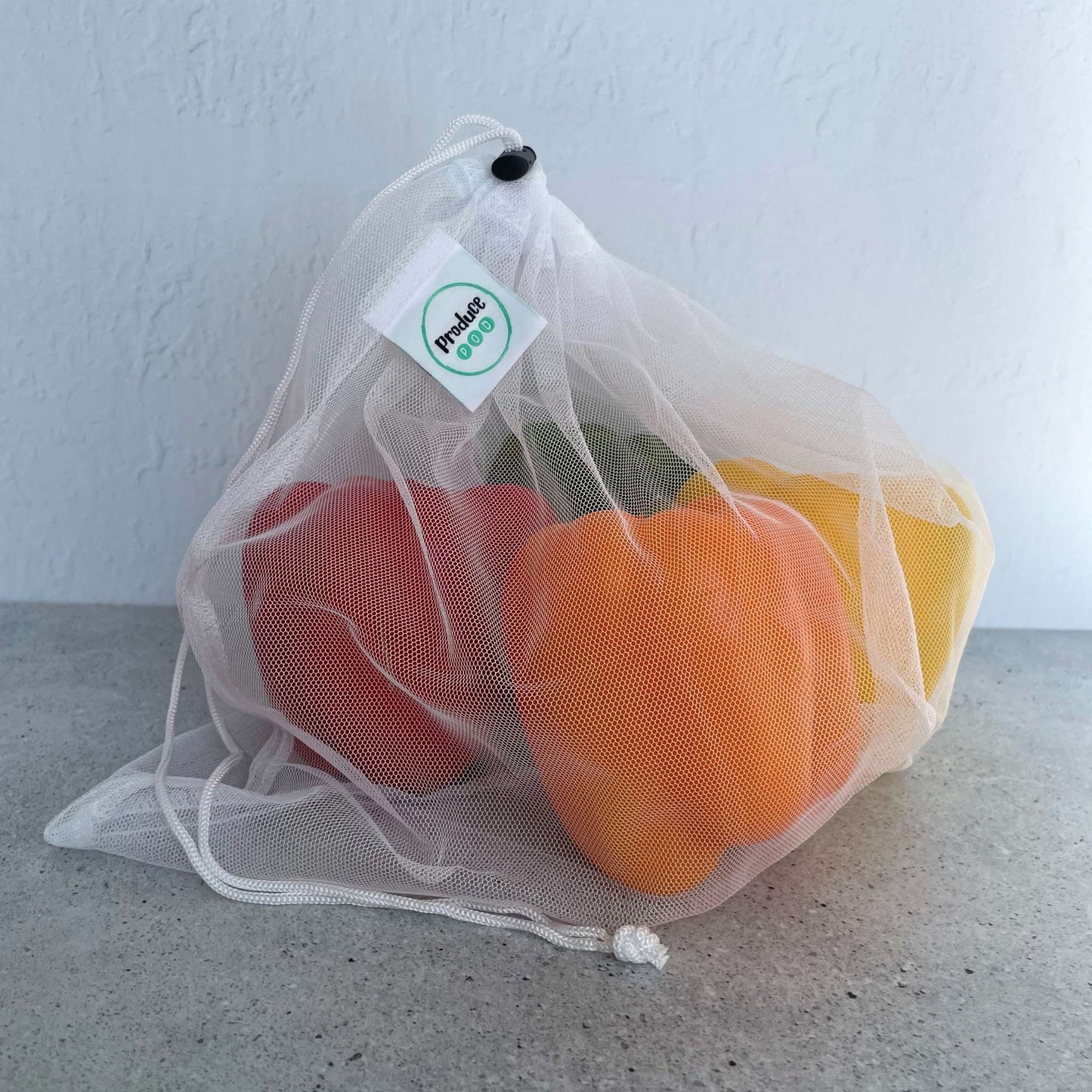 Mesh produce bag with red, orange, and yellow bell peppers on a gray surface.