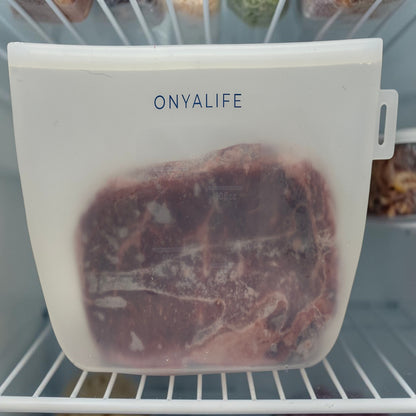 Reusable silicone food storage bag with flat base, pinch-seal top and slide-on sealing bar, showing measurement markings on translucent silicone. Small size shown inside freezer with a steak.