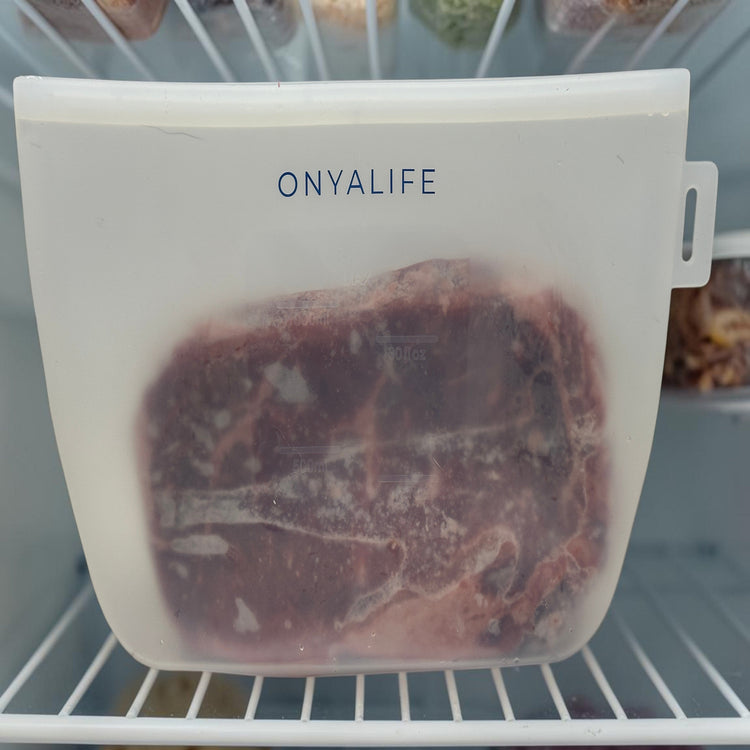 Reusable silicone food storage bag with flat base, pinch-seal top and slide-on sealing bar, showing measurement markings on translucent silicone. Small size shown inside freezer with a steak.