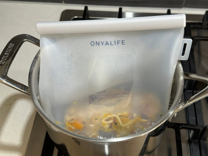 Reusable silicone food storage bag with flat base, pinch-seal top and slide-on sealing bar, showing measurement markings on translucent silicone. Shown immersed in hot water.