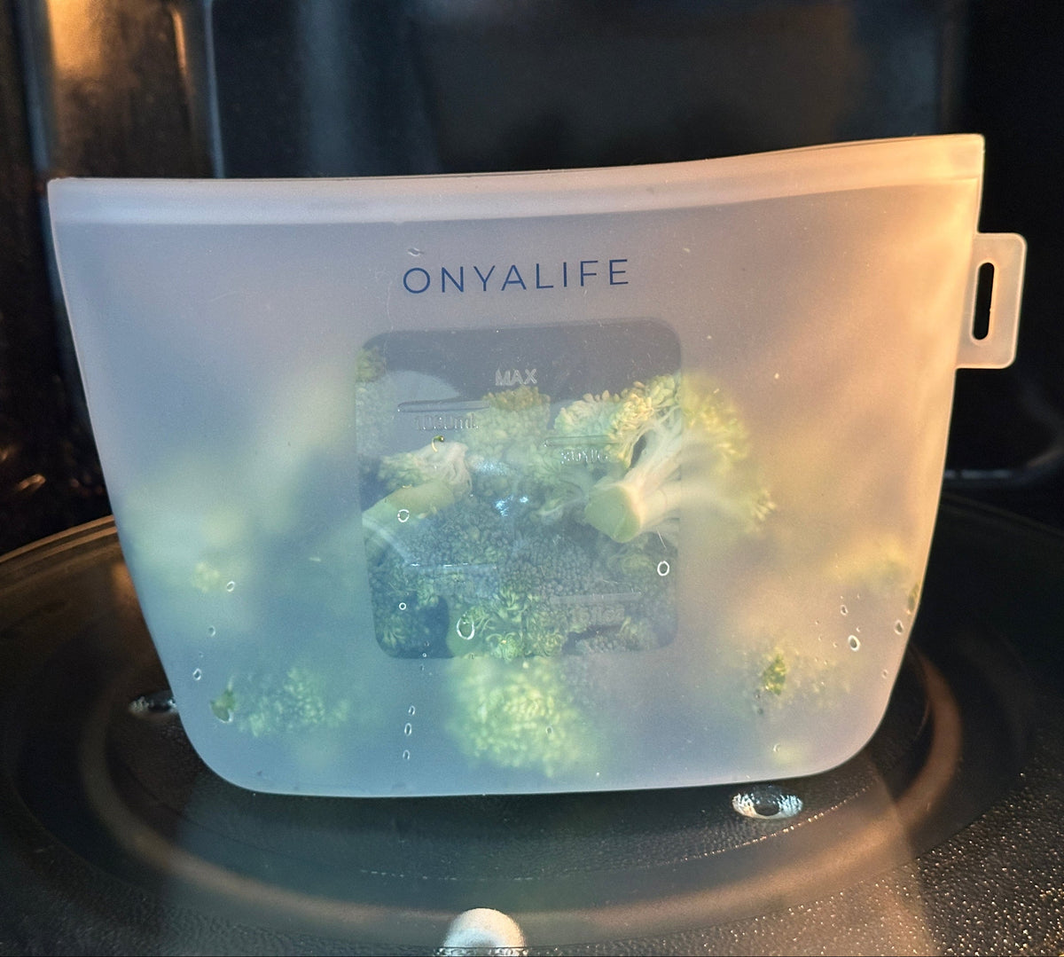 Reusable silicone food storage bag with flat base, pinch-seal top and slide-on sealing bar, showing measurement markings on translucent silicone. Shown inside microwave with broccoli