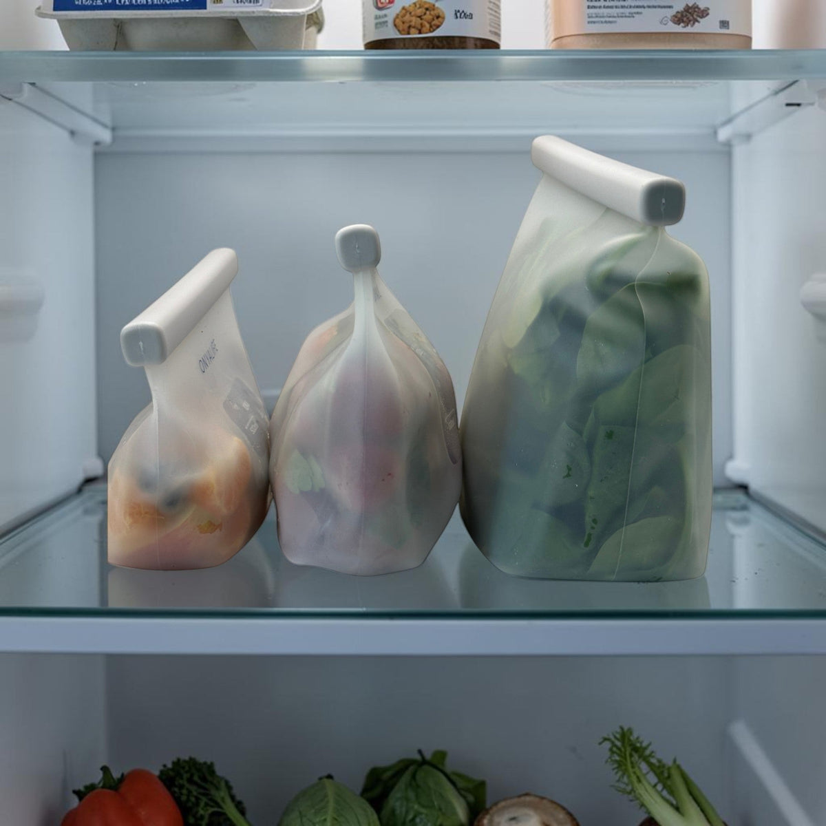 Reusable silicone food storage bag with flat base, pinch-seal top and slide-on sealing bar, showing measurement markings on translucent silicone. Shown in all 3 sizes on a shelf inside fridge.