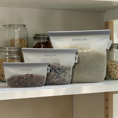 Reusable silicone food storage bag with flat base, pinch-seal top and slide-on sealing bar, showing measurement markings on translucent silicone. Shown in 3 sizes in pantry with dry foods