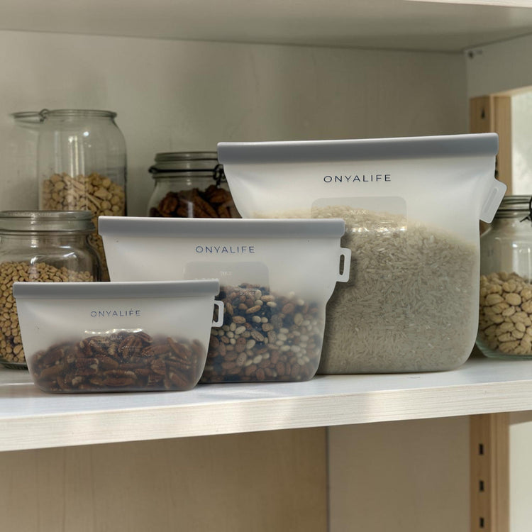 Reusable silicone food storage bag with flat base, pinch-seal top and slide-on sealing bar, showing measurement markings on translucent silicone. Shown in 3 sizes in pantry with dry foods