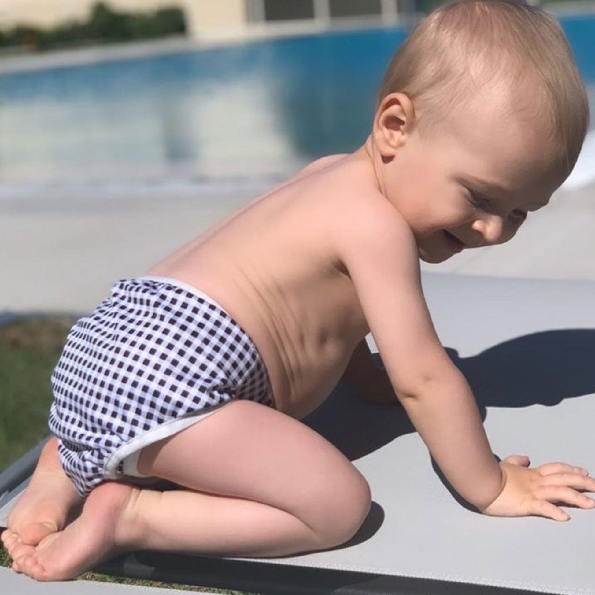 Happy toddler wearing a Tyoub black check reusable swim nappy by an outdoor pool in Australia.