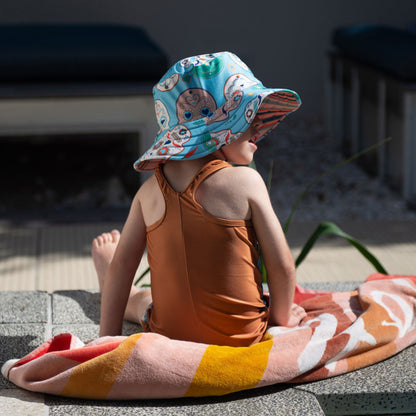 Child wearing the Tyoub sugar skull kneesuit zootie and matching sun hat sitting by an outdoor pool in Australia back view.