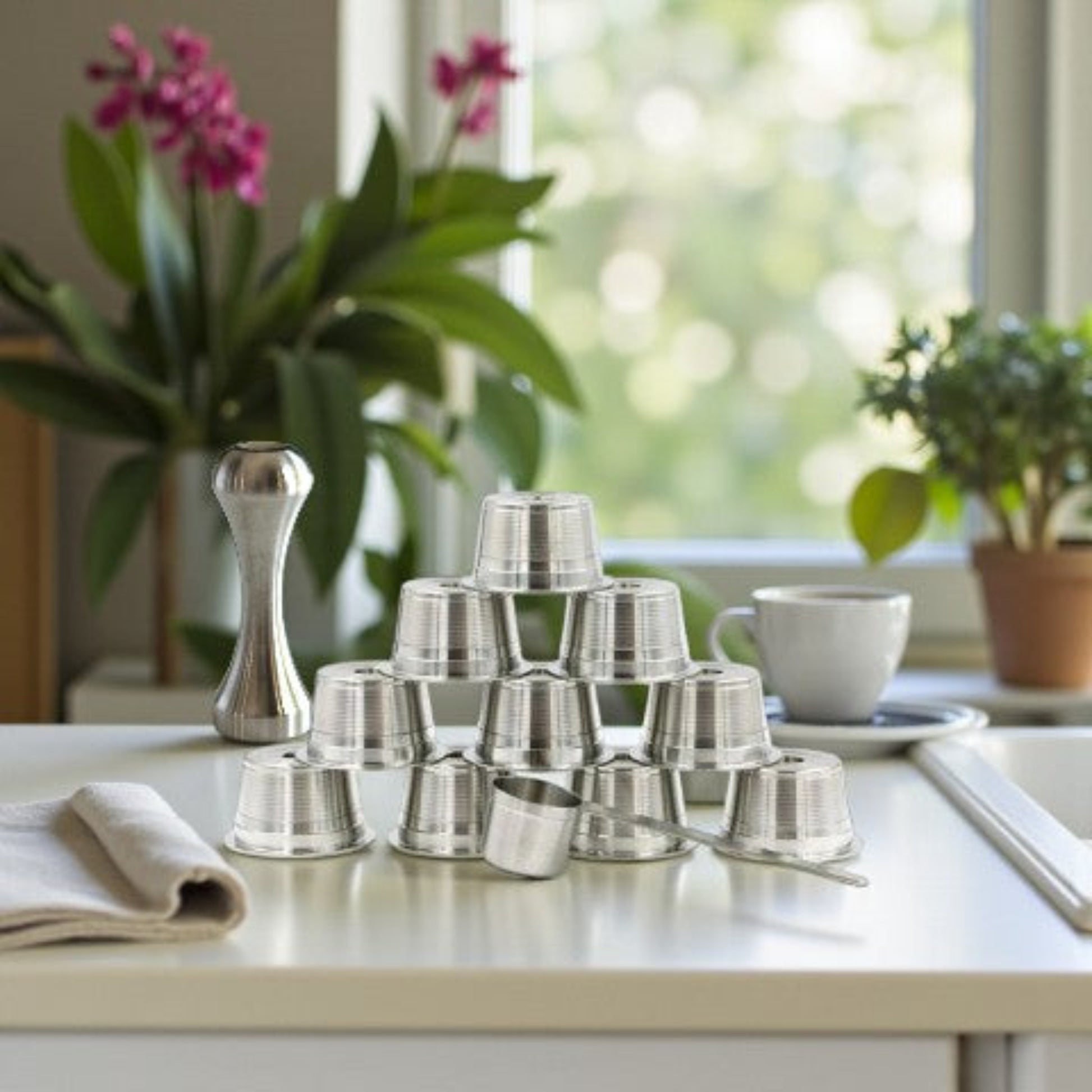 Ten eco-friendly stainless steel refillable coffee pods with a measuring scoop and espresso packing tamper, on a kitchen benchtop near a window.
