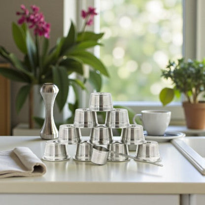 Ten eco-friendly stainless steel refillable coffee pods with a measuring scoop and espresso packing tamper, on a kitchen benchtop near a window.