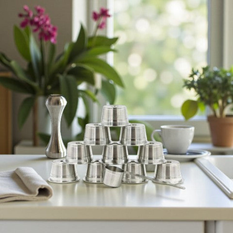 Ten eco-friendly stainless steel refillable coffee pods with a measuring scoop and espresso packing tamper, on a kitchen benchtop near a window.