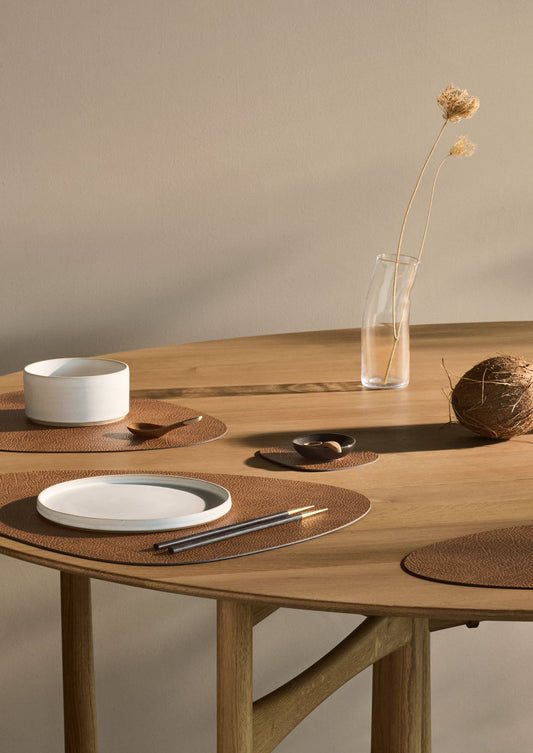 Recycled leather tablemats arranged on wooden dining table with ceramic plates, a white bowl, and a vase with a single dried flower on a neutral background.