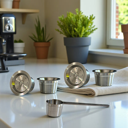 Four single stainless steel reusable coffee pods and a scoop next to a pot plant on a kitchen countertop with a napkin.