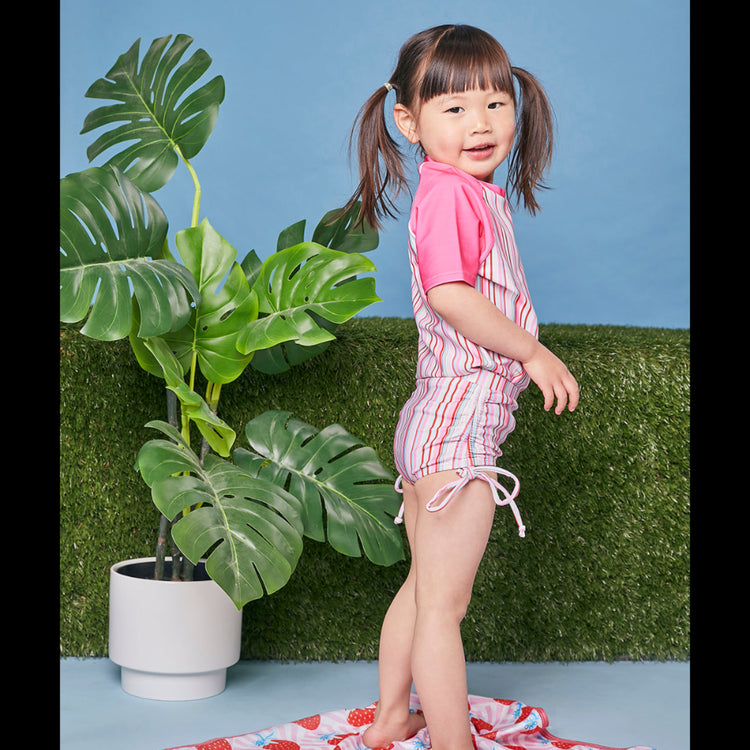 Young girl modeling the Tyoub short sleeve gelato pink striped rash vest with matching swimsuit side view.