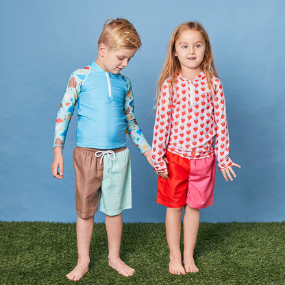 Young girl wearing the Tyoub strawberry sundae rash guard paired with pink swim board shorts in a studio setting.