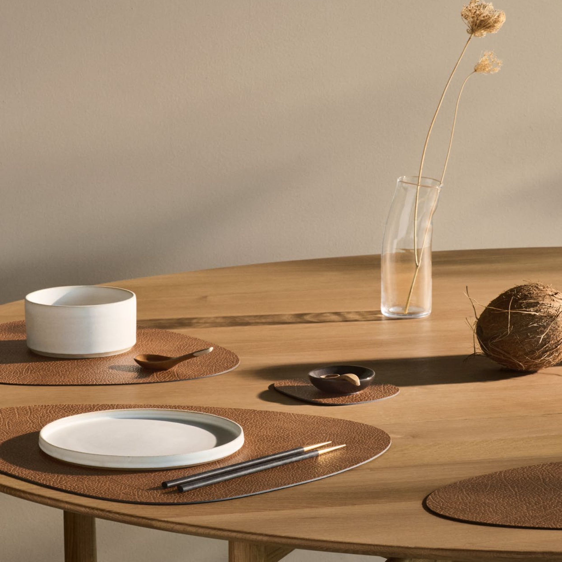 Curve recycled leather placemats featuring the signature LIND DNA hippo texture arranged on a wooden dining table with ceramic plates, a white bowl, and a vase with a single dried flower on a neutral background.