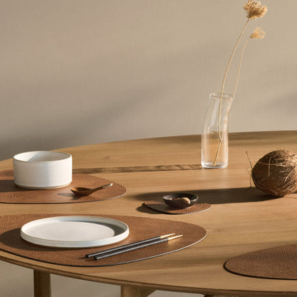 Curve recycled leather placemats featuring the signature LIND DNA hippo texture arranged on a wooden dining table with ceramic plates, a white bowl, and a vase with a single dried flower on a neutral background.