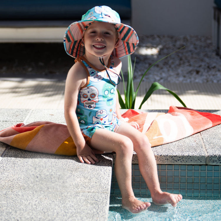 Child wearing the Tyoub sugar skull kneesuit zootie and matching sun hat sitting by an outdoor pool in Australia.