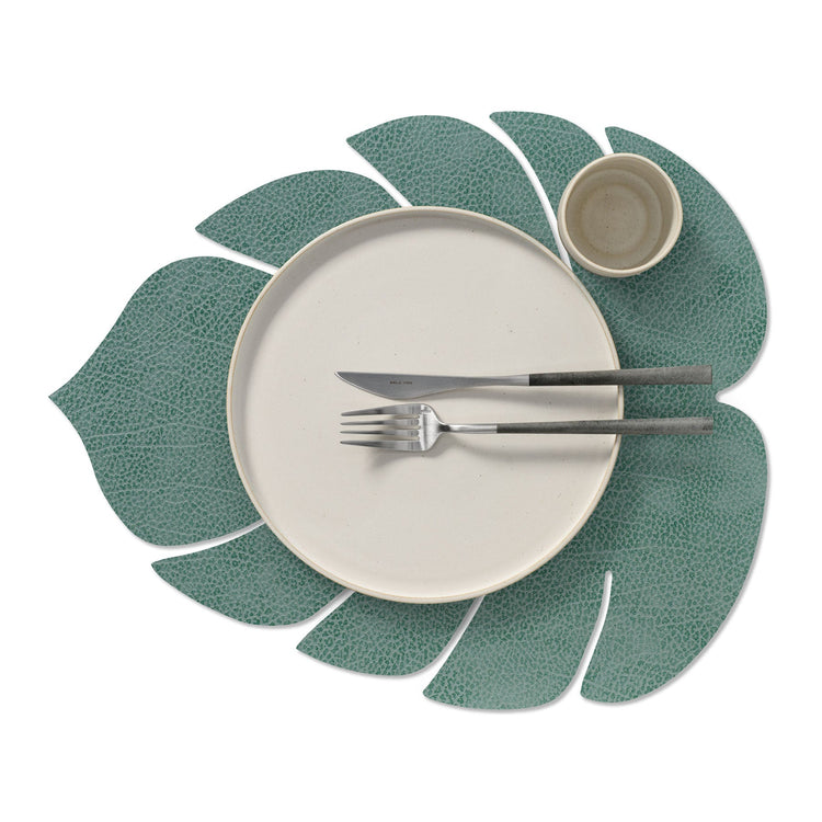 Monstera leaf recycled leather placemat featuring the signature LIND DNA Hippo finish in pastel green with a plate, cup, knife and fork.
