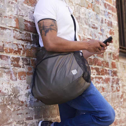 Man leaning against a brick wall while carrying a iightweight Onya packable crossbody side messenger bag showing an adjustable shoulder strap and spacious front compartment panel in Charcoal.