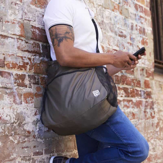 Man leaning against a brick wall while carrying a iightweight Onya packable crossbody side messenger bag showing an adjustable shoulder strap and spacious front compartment panel in Charcoal.