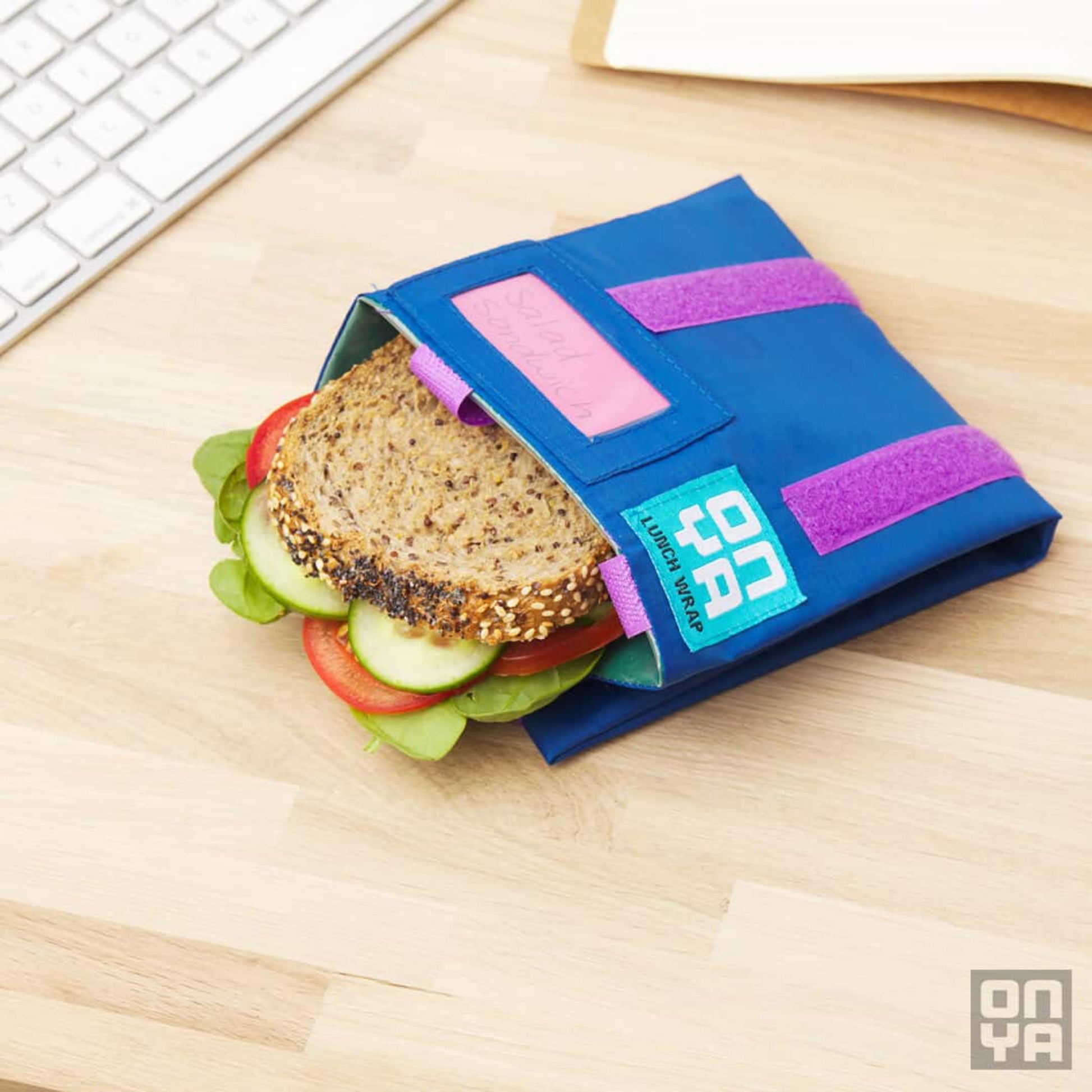 ONYA reusable lunch wrap, made from recycled rPeT  with a food safe PEVA lining. BPA free. Vegan friendly. Fully recycable at end of life. Showing how to use with a snadwich on a table top.
