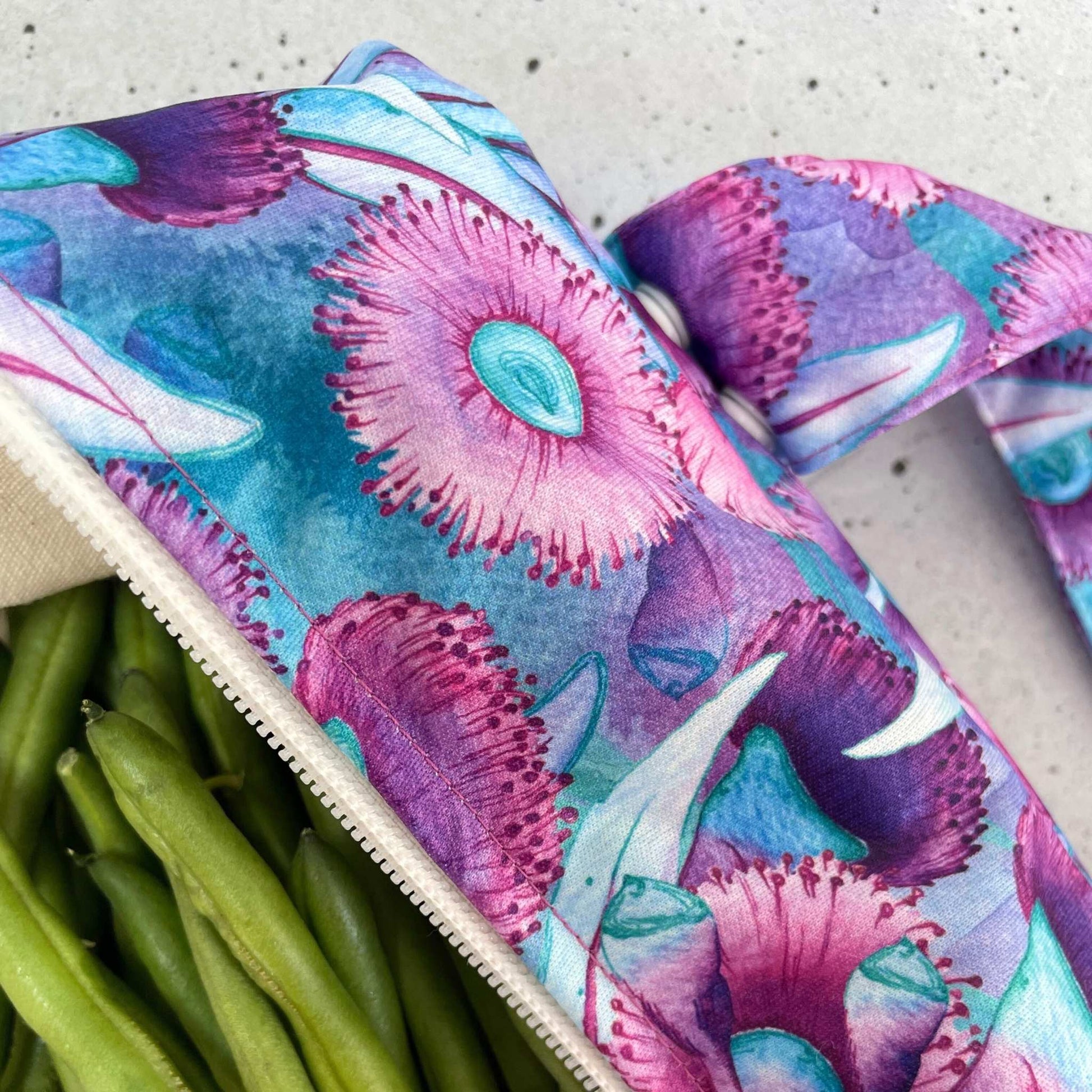 Single Produce Pod reusable veggie storage bag featuring a custom Gum blossom print standing open filled with fresh veg and fruit closeup with beans on a stone background.