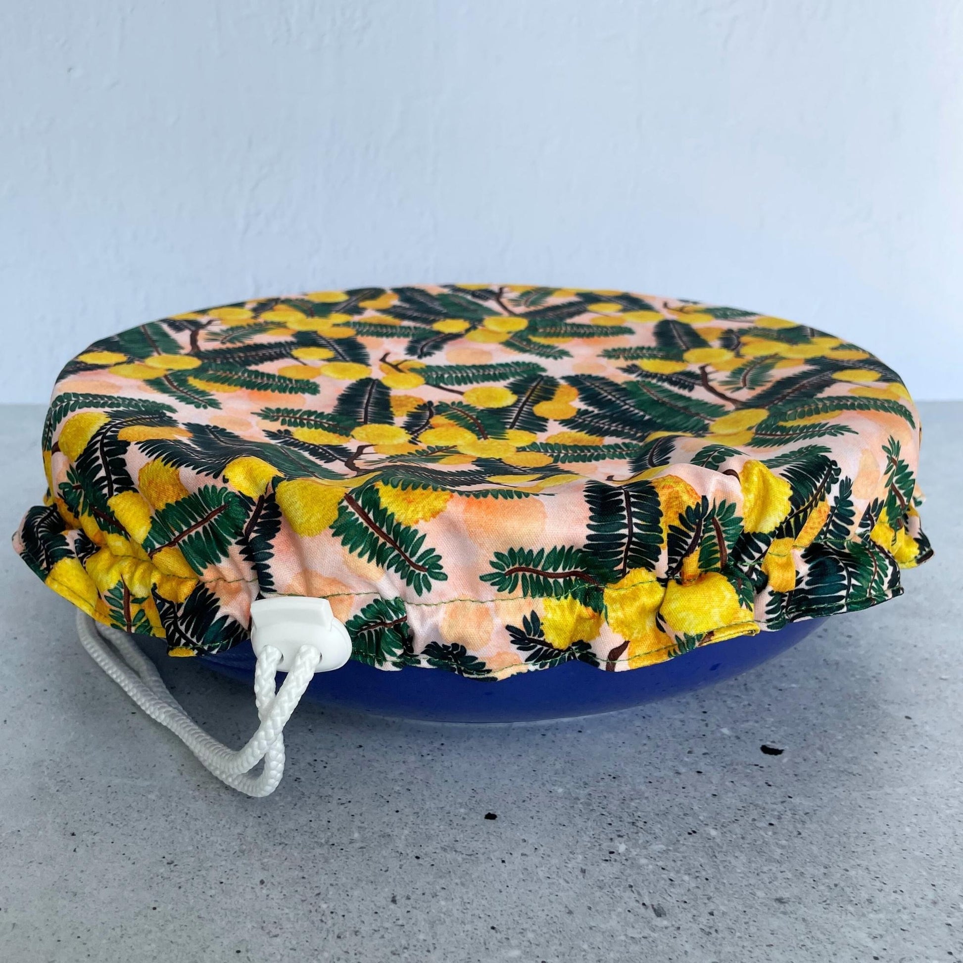 Produce Pod reusable bowl cover in a Wattle print fitted snugly over a blue serving dish on sone benchtop.