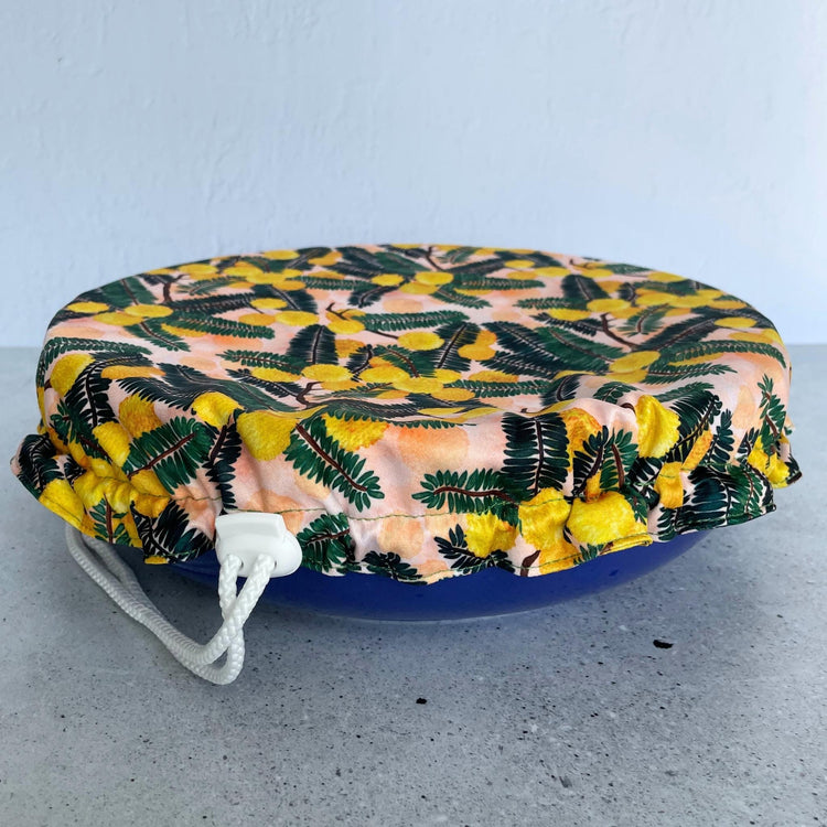 Produce Pod reusable bowl cover in a Wattle print fitted snugly over a blue serving dish on sone benchtop.