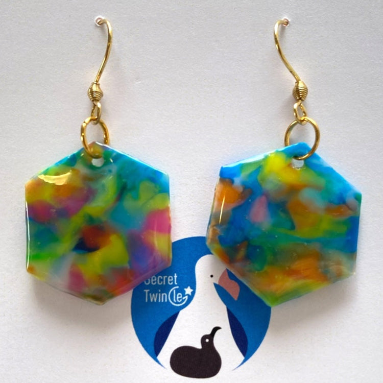 A pair of handmade recycled bottle cap statement earrings in a geometric hexagon dangle design with blue multi color marbling.