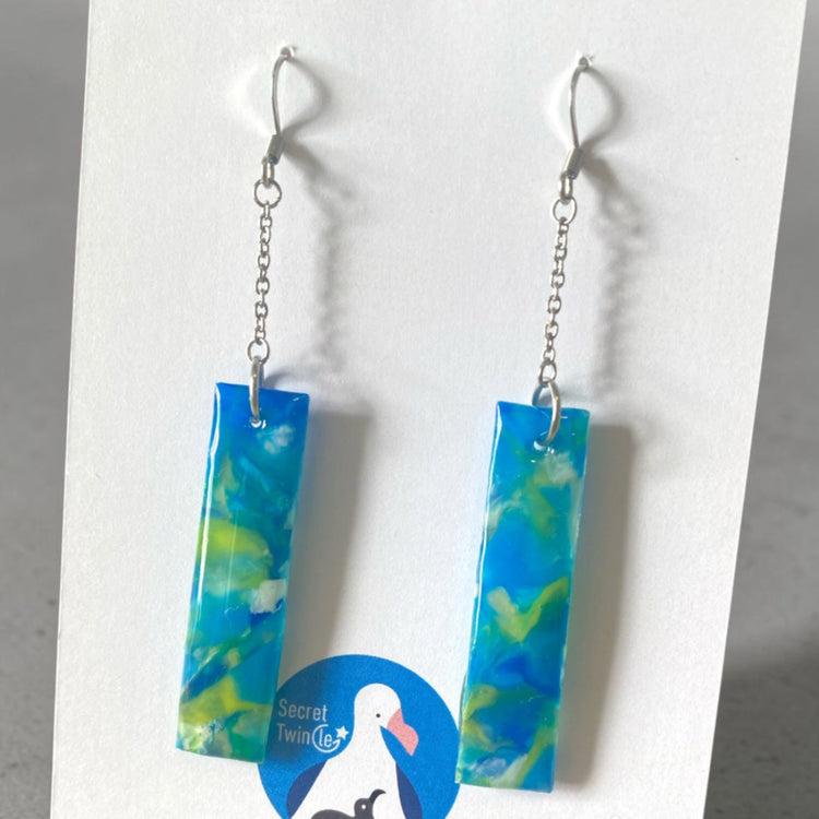 A pair of handmade upcycled plastic statement earrings featuring a geometric rectangular chain link dangle design in marbled blue and green tones.