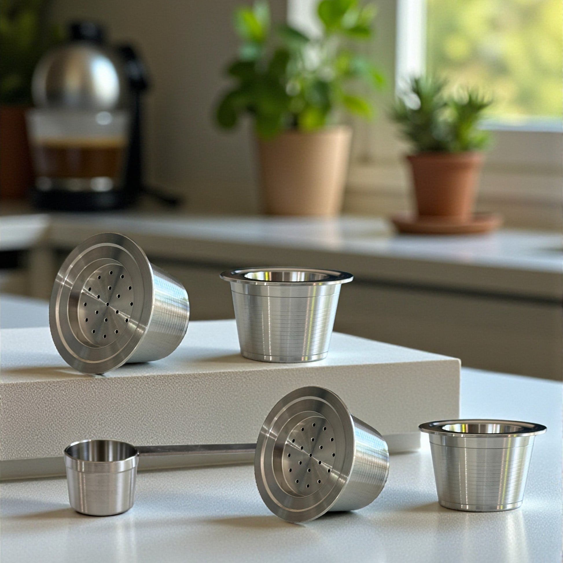 Four eco-friendly reusable stainless steel espresso capsules with a scoop arranged on a kithen benchtop near a window.
