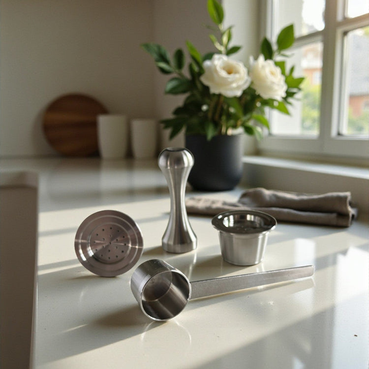 Two refillable stainless steel coffee pods displayed alongside a precision tamper and measuring scoop on a kitchen countertop near a window.