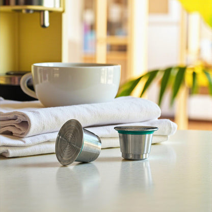 Two refillable stainless steel coffee pods on a kitchen countertop with a cup of coffee.