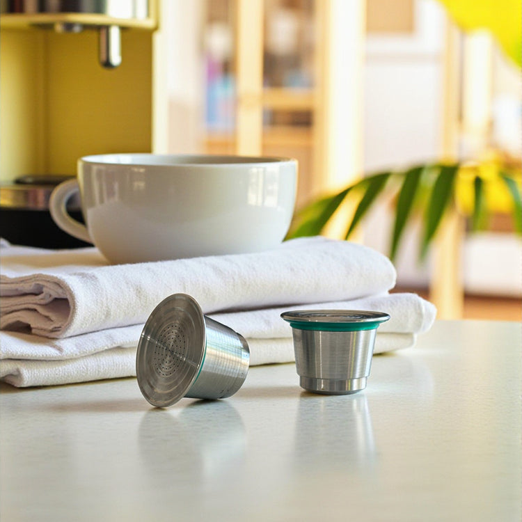 Two refillable stainless steel coffee pods on a kitchen countertop with a cup of coffee.