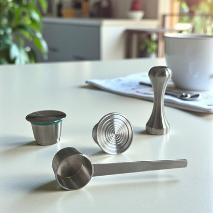 Two refillable stainless steel coffee pods displayed alongside a precision packing tamper and measuring scoop spoon on a kitchen countertop.