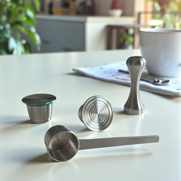 Two refillable stainless steel coffee pods displayed alongside a precision packing tamper and measuring scoop spoon on a kitchen countertop.