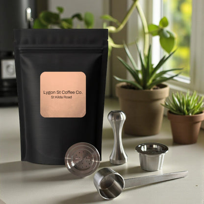 Pod Star starter pack for Espressotoria coffee machines featuring two refillable stainless steel capsules, an espresso tamper, a measuring scoop, and fresh coffee grounds on a kitchen countertop.