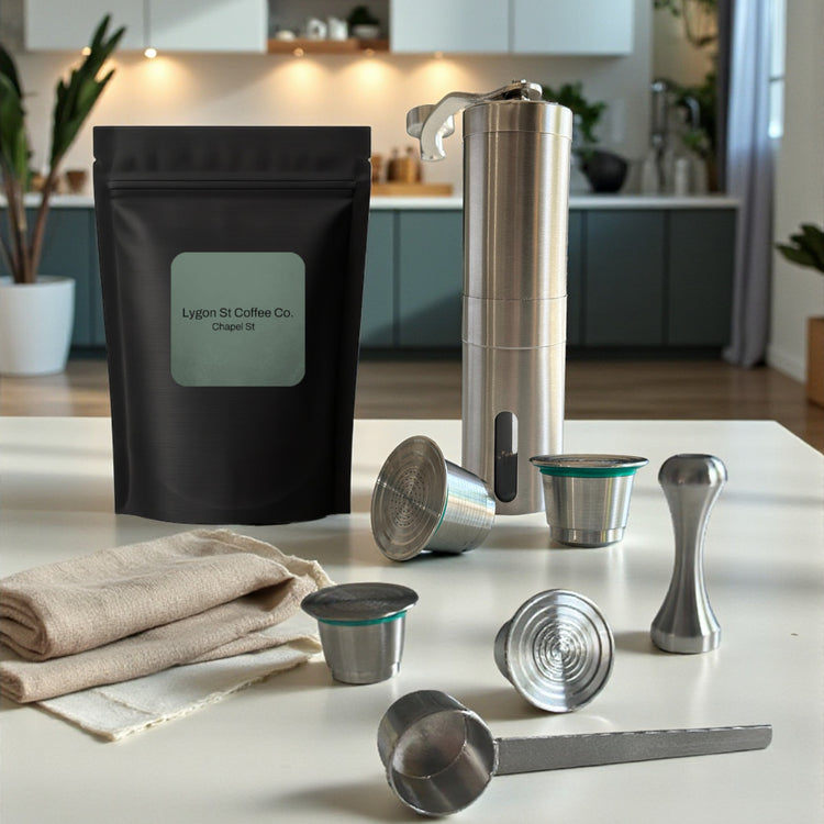 Pod Star ultimate bundle for Nespresso Original line coffee machines featuring four stainless steel reusable capsules, a manual hand grinder, an espresso tamper, and a coffee measuring scoop on a kitchen countertop.