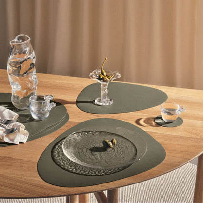 Recycled leather tablemats in Moss on adining table with placemats, glasses, and a vase against a beige curtain background