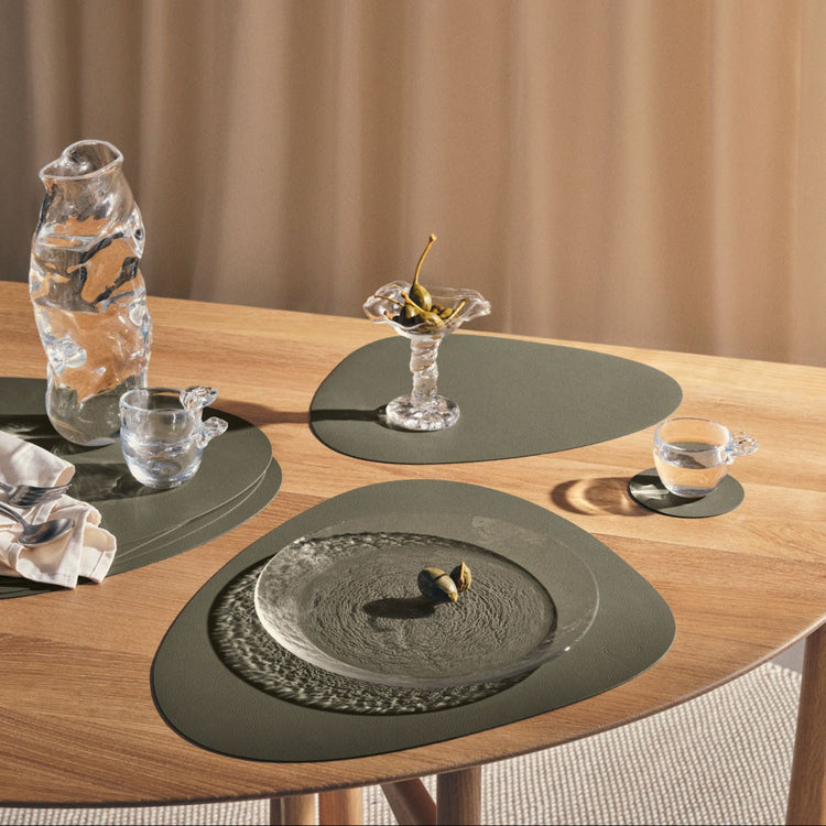 Recycled leather tablemats in Moss on adining table with placemats, glasses, and a vase against a beige curtain background