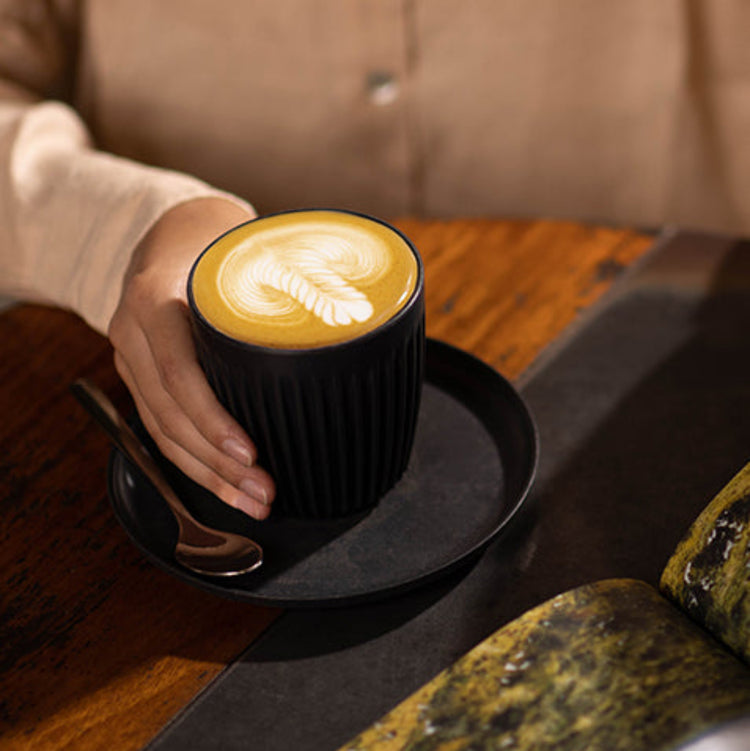 A durable Charcoal Huskee classic universal saucer styled beneath a matching reusable coffee cup on a kitchen table.