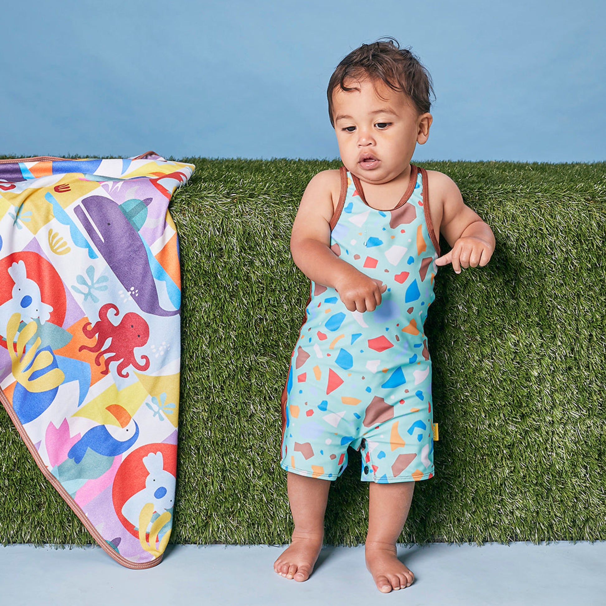 Toddler wearing the Tyoub ocean glass color block zootie swimsuit standing by a colorful baby beach rug.