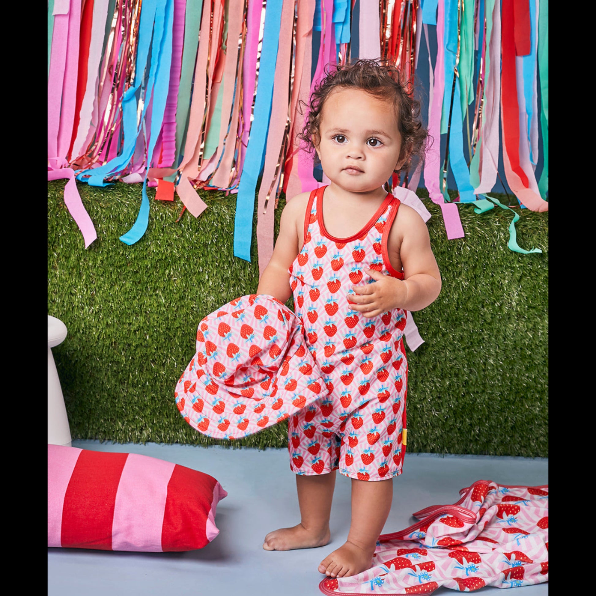 Young girl wearing a Tyoub strawberry sundae print zootie swimsuit holding a matching hat.