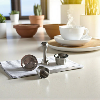 Two single stainless steel reusable coffee pods, tamper and scoop next to a cup of coffee and plate on a countertop.