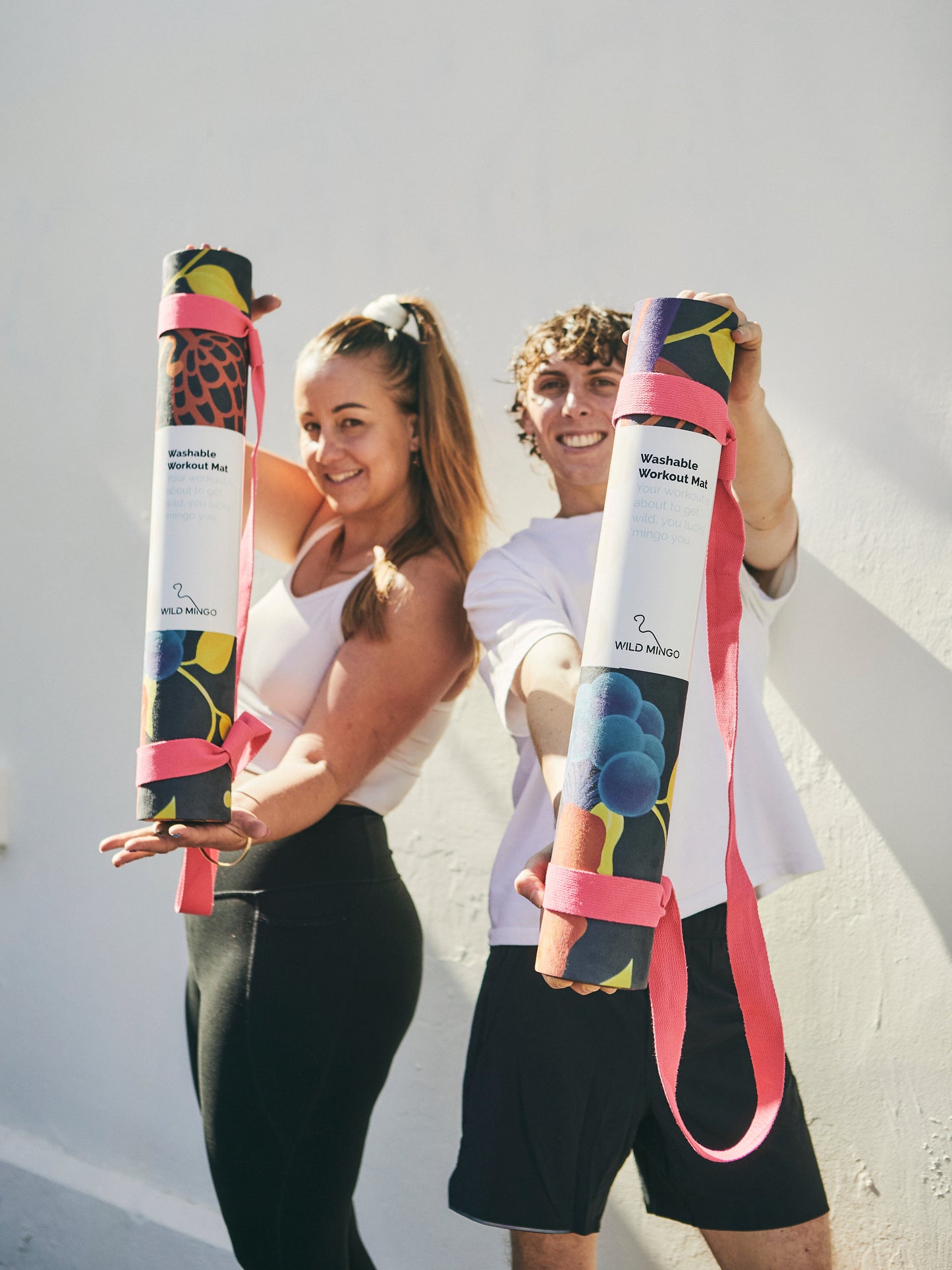 Two people holding rolled Wild Mingo Pink Lady Yoga Mats with a white stucco wall behind
